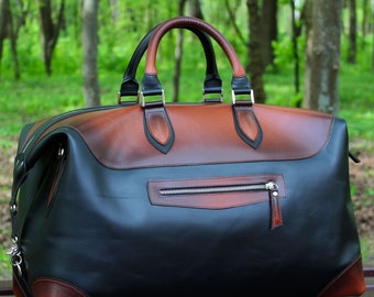 May include: A large duffel bag featuring a two-tone design with a smooth black leather body and burnished brown leather accents on the top panel, corners, and handles. The bag includes a horizontal zippered pocket on the front.