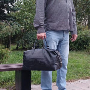 May include: A black duffel bag with a textured crocodile-skin pattern, featuring two top handles, silver-toned metal hardware, and a zippered closure. The bag has a rectangular shape with rounded corners and a structured base.