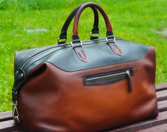 May include: A large duffel bag featuring a two-tone design with a textured brown leather body and a smooth dark green leather top panel. The bag includes two top handles, a zippered exterior pocket, and silver-toned metal hardware.