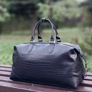 May include: A black duffel bag featuring a textured crocodile skin pattern. The bag has two top handles secured with a central snap closure, a horizontal zipper across the top, and silver-toned metal hardware attachments on the sides.