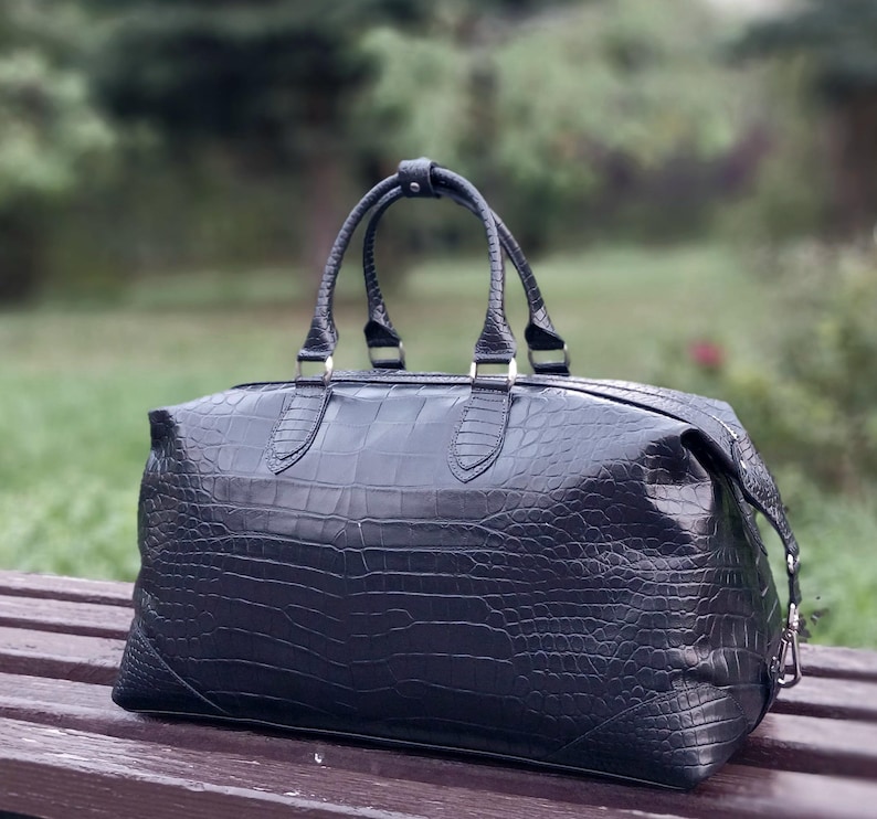May include: A black duffel bag featuring a textured crocodile skin pattern. The bag has two top handles secured with a central snap closure, a horizontal zipper across the top, and silver-toned metal hardware attachments on the sides.