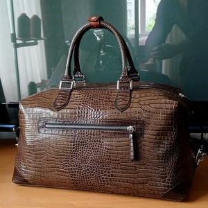 Puede incluir: Una bolsa de viaje de cuero marr&oacute;n con un patr&oacute;n de estampado de cocodrilo. La bolsa tiene dos asas superiores y una correa de hombro desmontable.