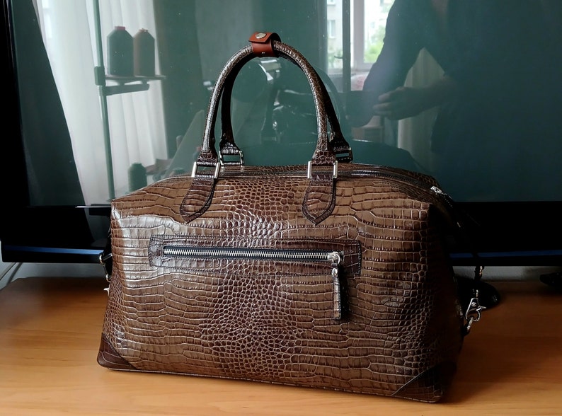 Puede incluir: Una bolsa de viaje de cuero marr&oacute;n con un patr&oacute;n de estampado de cocodrilo. La bolsa tiene dos asas superiores y una correa de hombro desmontable.
