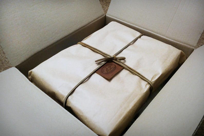 May include: A rectangular package wrapped in light brown paper sits inside an open cardboard box. The package is secured with a thin brown cord tied in a bow, featuring a small, square, dark brown leather tag with an embossed circular logo.