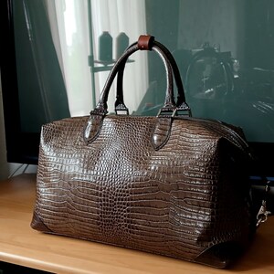 Puede incluir: Una bolsa de viaje de cuero marr&oacute;n con estampado de cocodrilo, con asa de cuero marr&oacute;n y cierre de cremallera.
