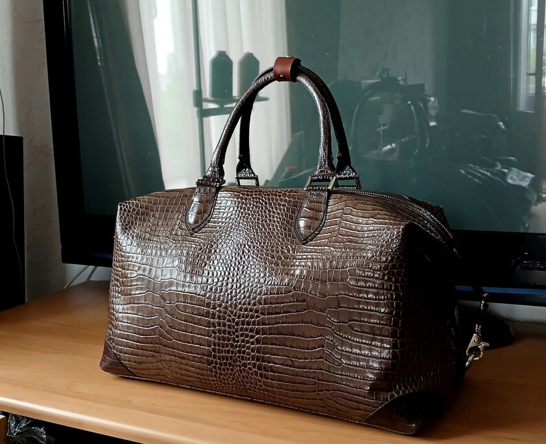 Puede incluir: Una bolsa de viaje de cuero marr&oacute;n con estampado de cocodrilo, con asa de cuero marr&oacute;n y cierre de cremallera.