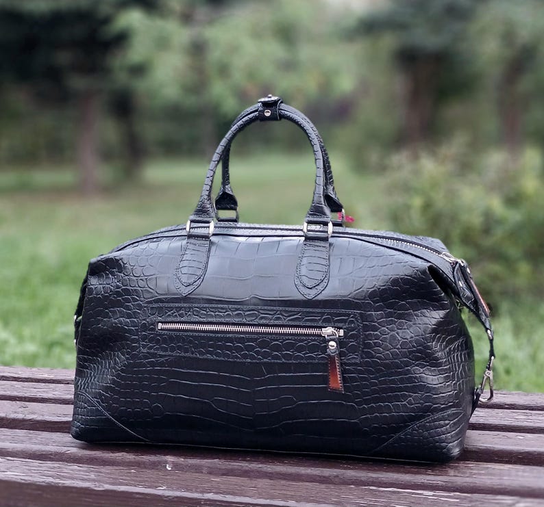 May include: A black duffel bag with a textured crocodile skin pattern. The bag features two top handles, a silver-toned horizontal zipper pocket on the front, and a detachable shoulder strap with silver metal hardware clips on the sides.