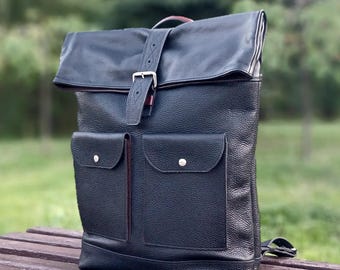 Pode incluir: Uma mochila de couro preta com fecho de enrolar, acabamento granulado texturizado, dois bolsos frontais com fechos de pressão prateados e uma tira de couro vertical com fivela de metal para prender a aba. O design inclui uma alça superior.