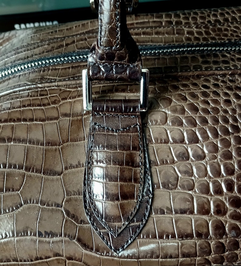 Puede incluir: Primer plano de un bolso de mano de cuero de cocodrilo marr&oacute;n con una hebilla plateada y un asa superior.