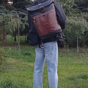 Pode incluir: Uma mochila roll-top com um painel frontal de couro castanho escuro com textura de crocodilo, contrastado com sec&ccedil;&otilde;es de couro preto liso na parte superior e na base. O saco inclui uma fivela de metal prateado e um bolso com fecho.