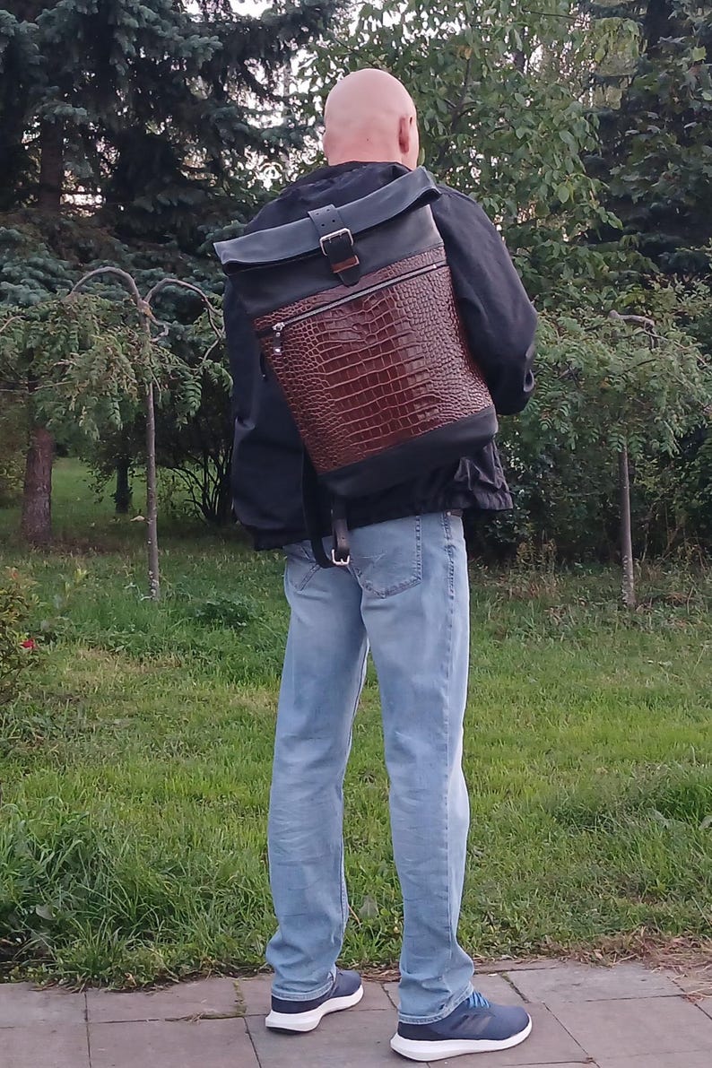 Pode incluir: Uma mochila roll-top com um painel frontal de couro castanho escuro com textura de crocodilo, contrastado com sec&ccedil;&otilde;es de couro preto liso na parte superior e na base. O saco inclui uma fivela de metal prateado e um bolso com fecho.