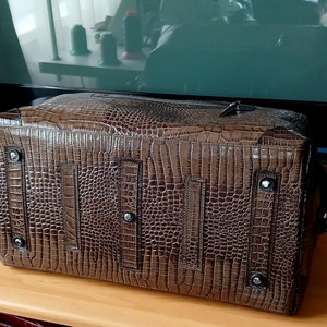 Puede incluir: Una bolsa de viaje de cuero marr&oacute;n con un patr&oacute;n de piel de cocodrilo. La bolsa tiene una cremallera y dos correas en la parte posterior.