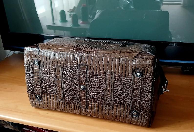 Puede incluir: Una bolsa de viaje de cuero marr&oacute;n con un patr&oacute;n de piel de cocodrilo. La bolsa tiene una cremallera y dos correas en la parte posterior.