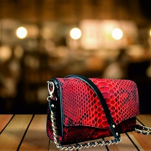 May include: A red snakeskin crossbody bag with a black strap and gold chain. The bag has a flap closure and a small pocket on the front.
