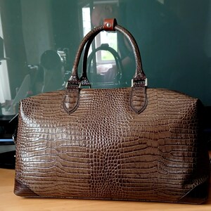 Puede incluir: Una bolsa de viaje de cuero marr&oacute;n con estampado de cocodrilo. La bolsa tiene un asa superior y una correa para el hombro.