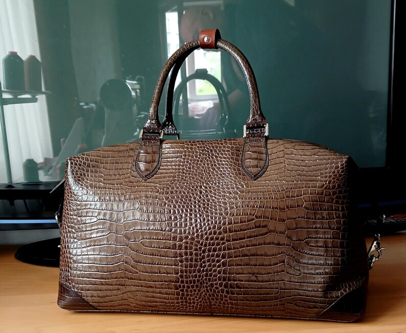 Puede incluir: Una bolsa de viaje de cuero marr&oacute;n con estampado de cocodrilo. La bolsa tiene un asa superior y una correa para el hombro.