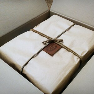 Puede incluir: Una caja de cart&oacute;n marr&oacute;n con un paquete envuelto en papel blanco en su interior. El paquete est&aacute; atado con cuerda y tiene una etiqueta de cuero marr&oacute;n con la palabra "12" en ella.