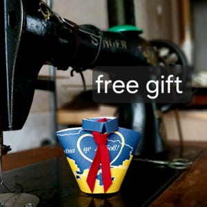 May include: A paper craft shirt ornament featuring a blue and yellow map design with a heart outline and the text "free gift" and "аїна це Любов!". The shirt has a small blue collar and a bright red ribbon tie centered on the front.