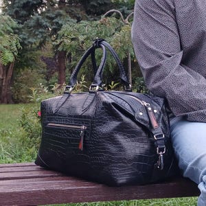 May include: A black travel bag with a textured crocodile skin pattern, featuring two top handles, a silver-toned metal zipper closure, and an exterior zippered pocket on the front. The bag has a rectangular shape with reinforced corners.