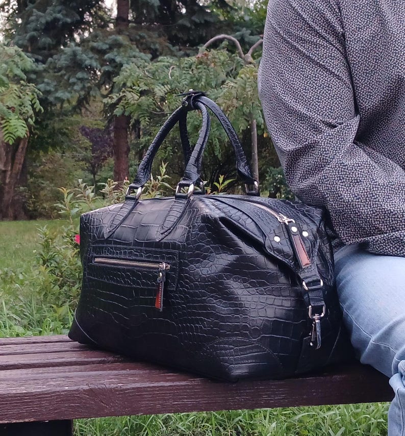 May include: A black travel bag with a textured crocodile skin pattern, featuring two top handles, a silver-toned metal zipper closure, and an exterior zippered pocket on the front. The bag has a rectangular shape with reinforced corners.