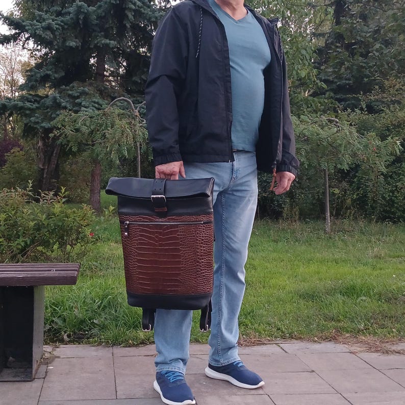 Pode incluir: Uma mochila roll-top com uma sec&ccedil;&atilde;o superior em pele preta e uma sec&ccedil;&atilde;o inferior em pele castanha com um padr&atilde;o texturado de pele de crocodilo. O saco inclui um bolso frontal com fecho, um fecho de fivela met&aacute;lica e al&ccedil;as pretas ajust&aacute;veis.