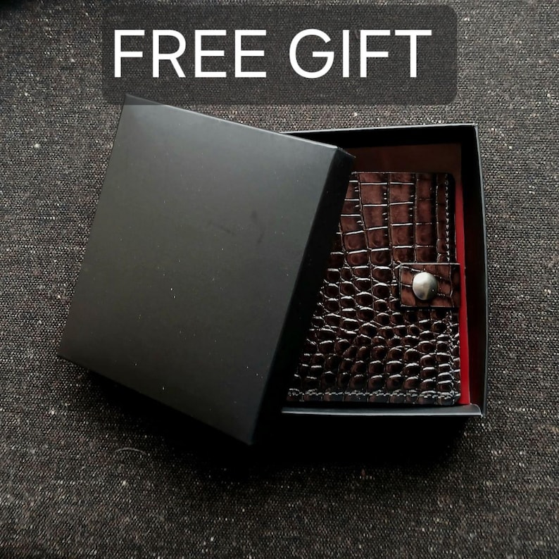 Pode incluir: Uma carteira castanho-escura com um padr&atilde;o texturizado de pele de crocodilo e um fecho de bot&atilde;o de press&atilde;o prateado, apresentada dentro de uma caixa de presente preta aberta. A tampa tem o texto FREE GIFT impresso em letras brancas.
