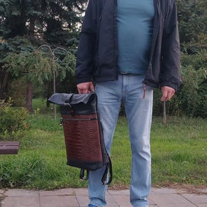 Pode incluir: Uma mochila de enrolar com parte superior e base em couro preto, apresentando um painel central em couro castanho texturizado com relevo de crocodilo. A bolsa inclui um fecho de fivela de metal prateado na aba e um bolso horizontal com z&iacute;per.