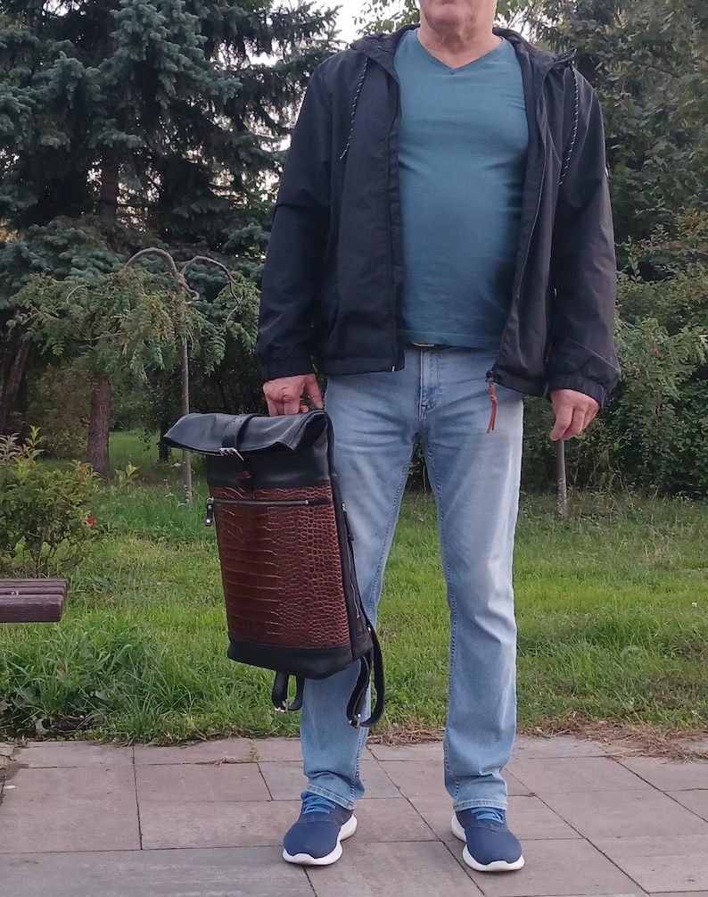 Pode incluir: Uma mochila de enrolar com parte superior e base em couro preto, apresentando um painel central em couro castanho texturizado com relevo de crocodilo. A bolsa inclui um fecho de fivela de metal prateado na aba e um bolso horizontal com z&iacute;per.