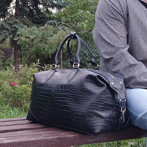 May include: A black duffel bag with a textured crocodile skin pattern, featuring two top handles, silver-toned metal hardware, and a zippered closure. The bag has a structured rectangular shape with reinforced corners and side attachments.
