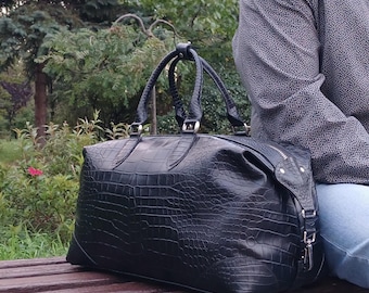 Könnte beinhalten: Eine schwarze Reisetasche mit texturiertem Krokodilleder-Muster, ausgestattet mit zwei Tragegriffen, silberfarbenen Metallbeschlägen und einem Reißverschluss. Die Tasche hat eine strukturierte rechteckige Form mit verstärkten Ecken.