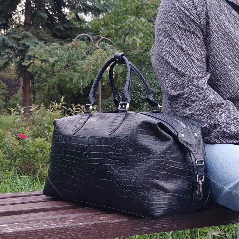 May include: A black duffel bag with a textured crocodile skin pattern, featuring two top handles, silver-toned metal hardware, and a zippered closure. The bag has a structured rectangular shape with reinforced corners and side attachments.