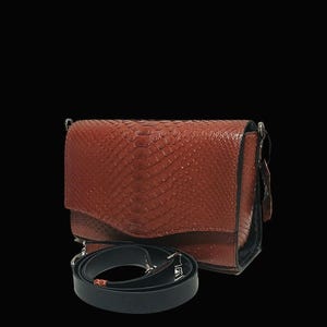 May include: A brown leather crossbody bag with a snakeskin texture. The bag has a black adjustable strap and silver hardware. The bag is rectangular with a flap closure and a black interior.