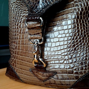 Puede incluir: Un bolso de mano de cuero marr&oacute;n con estampado de cocodrilo, con cierre de broche plateado y correa. El bolso tiene cierre de solapa y asa superior.