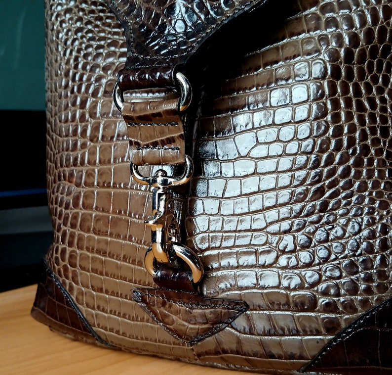 Puede incluir: Un bolso de mano de cuero marr&oacute;n con estampado de cocodrilo, con cierre de broche plateado y correa. El bolso tiene cierre de solapa y asa superior.