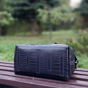 May include: A rectangular black handbag featuring a textured crocodile skin pattern. The bag has a structured design with vertical stitched panels along the side and a silver-toned metal hardware attachment on the right edge for a strap.