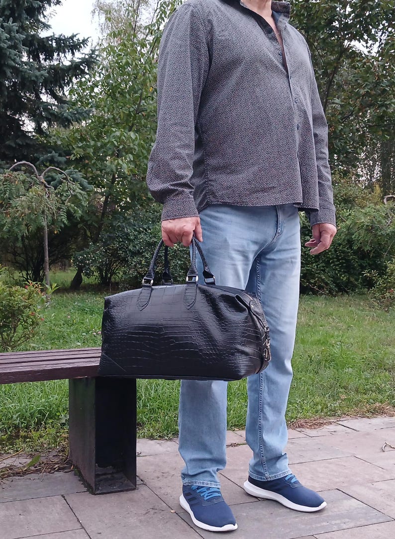 May include: A black duffel bag with a textured crocodile-skin pattern, featuring two top handles, silver-toned metal hardware, and a zippered closure. The bag has a rectangular shape with rounded corners and a structured base.