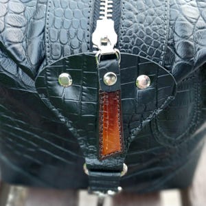 May include: A black bag with a textured crocodile-embossed pattern featuring a silver metal zipper and a triangular leather pull tab. The pull tab is black with a central brown gradient accent and is secured by three silver-toned metal rivets.