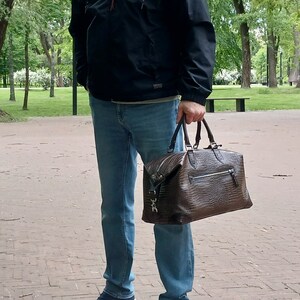 Puede incluir: Un hombre con una chaqueta negra, jeans azules y zapatillas azules lleva una bolsa de viaje de cuero marr&oacute;n con cremallera y hebilla plateada.