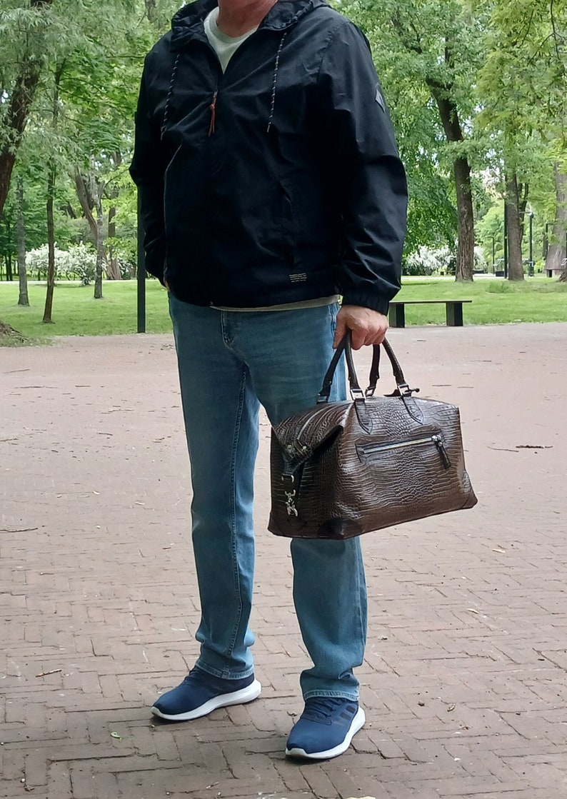 Puede incluir: Un hombre con una chaqueta negra, jeans azules y zapatillas azules lleva una bolsa de viaje de cuero marr&oacute;n con cremallera y hebilla plateada.
