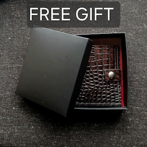 May include: A dark brown wallet with a textured crocodile-skin pattern and a silver-toned snap button closure, nestled inside an open black gift box. The box lid is partially covering the wallet, and the text FREE GIFT is visible.