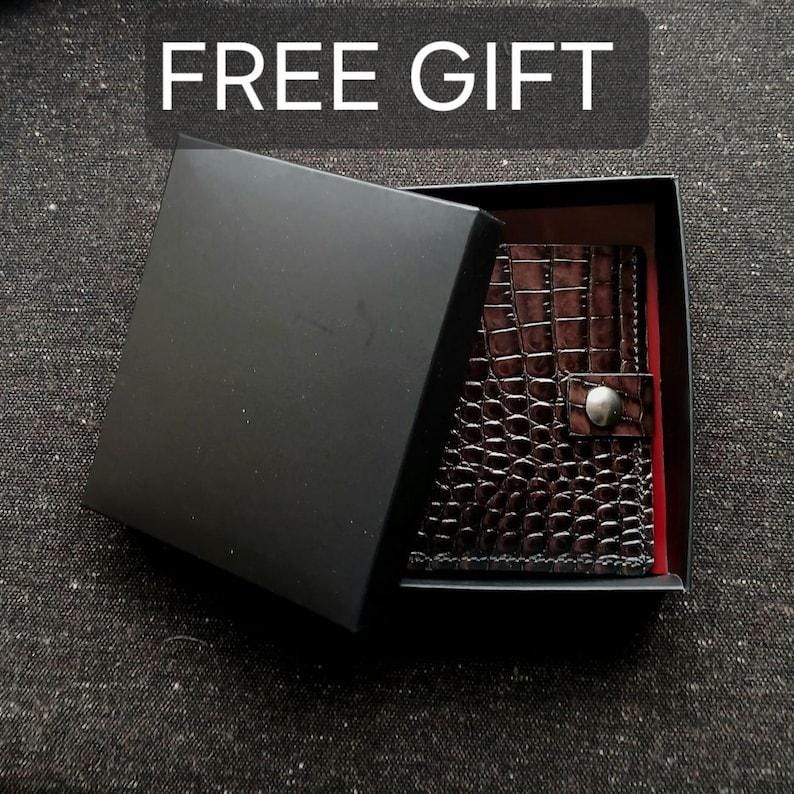 May include: A dark brown wallet with a textured crocodile-skin pattern and a silver-toned snap button closure, nestled inside an open black gift box. The box lid is partially covering the wallet, and the text FREE GIFT is visible.