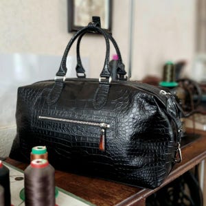 May include: A black duffel bag with a textured crocodile skin pattern, featuring two top handles, a horizontal zippered front pocket with a silver-toned metal zipper, and a small brown leather pull tab attached to the zipper slider.