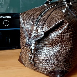 Puede incluir: Una bolsa de viaje de cuero marr&oacute;n con estampado de cocodrilo. La bolsa tiene cierre de cremallera y una hebilla de metal.