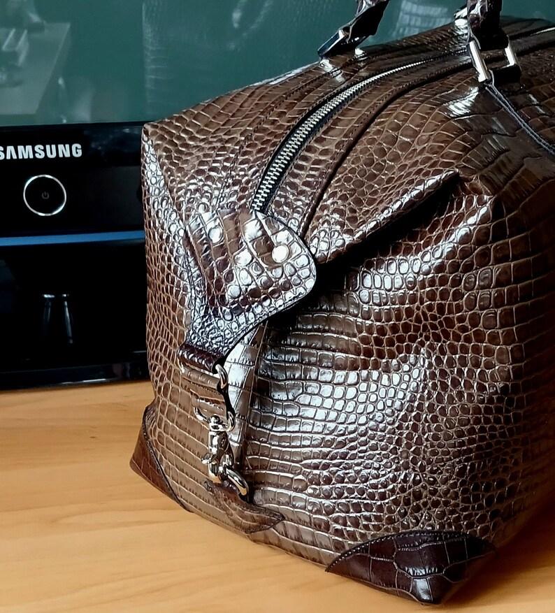 Puede incluir: Una bolsa de viaje de cuero marr&oacute;n con estampado de cocodrilo. La bolsa tiene cierre de cremallera y una hebilla de metal.
