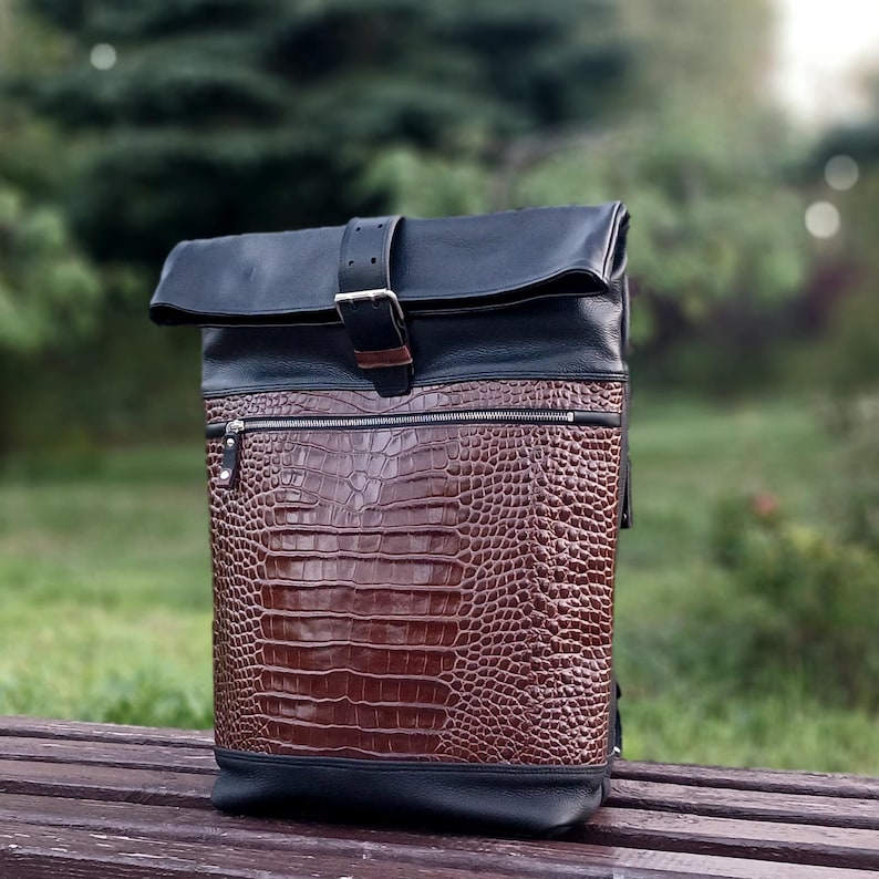 Pode incluir: Uma mochila roll-top com topo e base em couro preto e um painel frontal em couro castanho com relevo de crocodilo. A mala inclui um bolso horizontal com fecho na parte da frente e uma correia de couro preto com uma fivela de metal prateado.