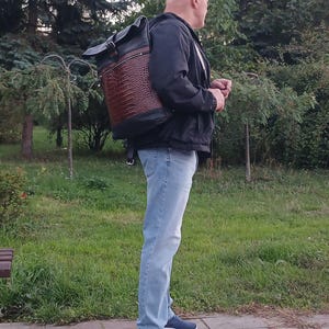 Pode incluir: Uma mochila roll-top com corpo em couro castanho com relevo de crocodilo, acabamentos em couro preto, fecho de fivela superior e um bolso horizontal com fecho de correr na frente, usada sobre um casaco preto, cal&ccedil;as de ganga azul-claras e sapatilhas azuis.