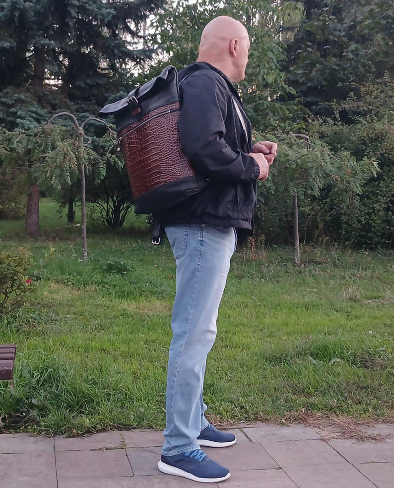 Pode incluir: Uma mochila roll-top com corpo em couro castanho com relevo de crocodilo, acabamentos em couro preto, fecho de fivela superior e um bolso horizontal com fecho de correr na frente, usada sobre um casaco preto, cal&ccedil;as de ganga azul-claras e sapatilhas azuis.