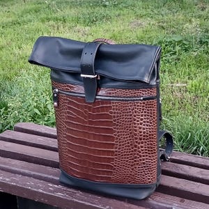Pode incluir: Uma mochila de fecho enrol&aacute;vel com uma sec&ccedil;&atilde;o superior em pele preta e um painel frontal em pele castanha com relevo de crocodilo. O saco inclui uma correia central vertical em pele preta com fivela de metal prateado e um bolso com fecho.