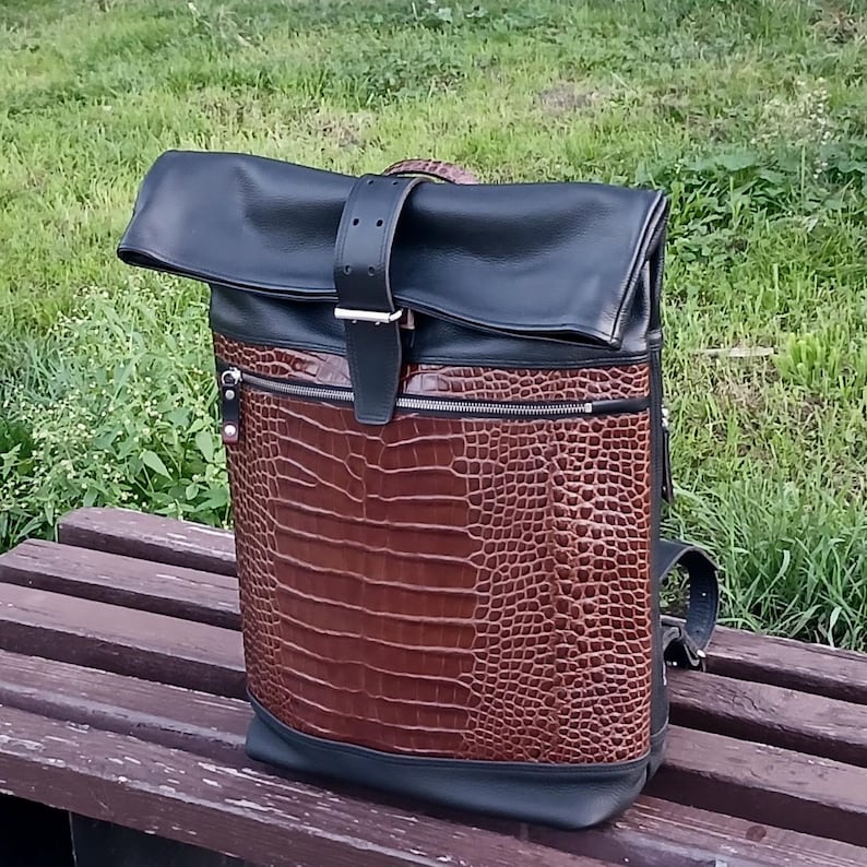 Pode incluir: Uma mochila de fecho enrol&aacute;vel com uma sec&ccedil;&atilde;o superior em pele preta e um painel frontal em pele castanha com relevo de crocodilo. O saco inclui uma correia central vertical em pele preta com fivela de metal prateado e um bolso com fecho.