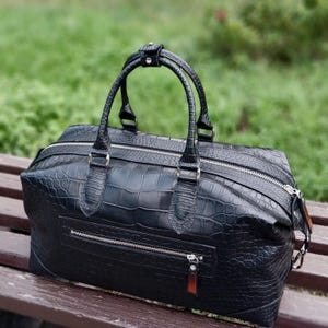 May include: A black duffel bag with a textured crocodile skin pattern, featuring two top handles, a main zipper closure, and a horizontal zippered pocket on the front. The bag has silver-toned hardware and a rectangular shape.